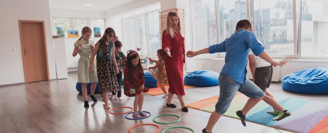 Small nursery school children with female teacher on floor indoors in classroom, doing exercise. Jumping over hula hoop circles track on the floor.