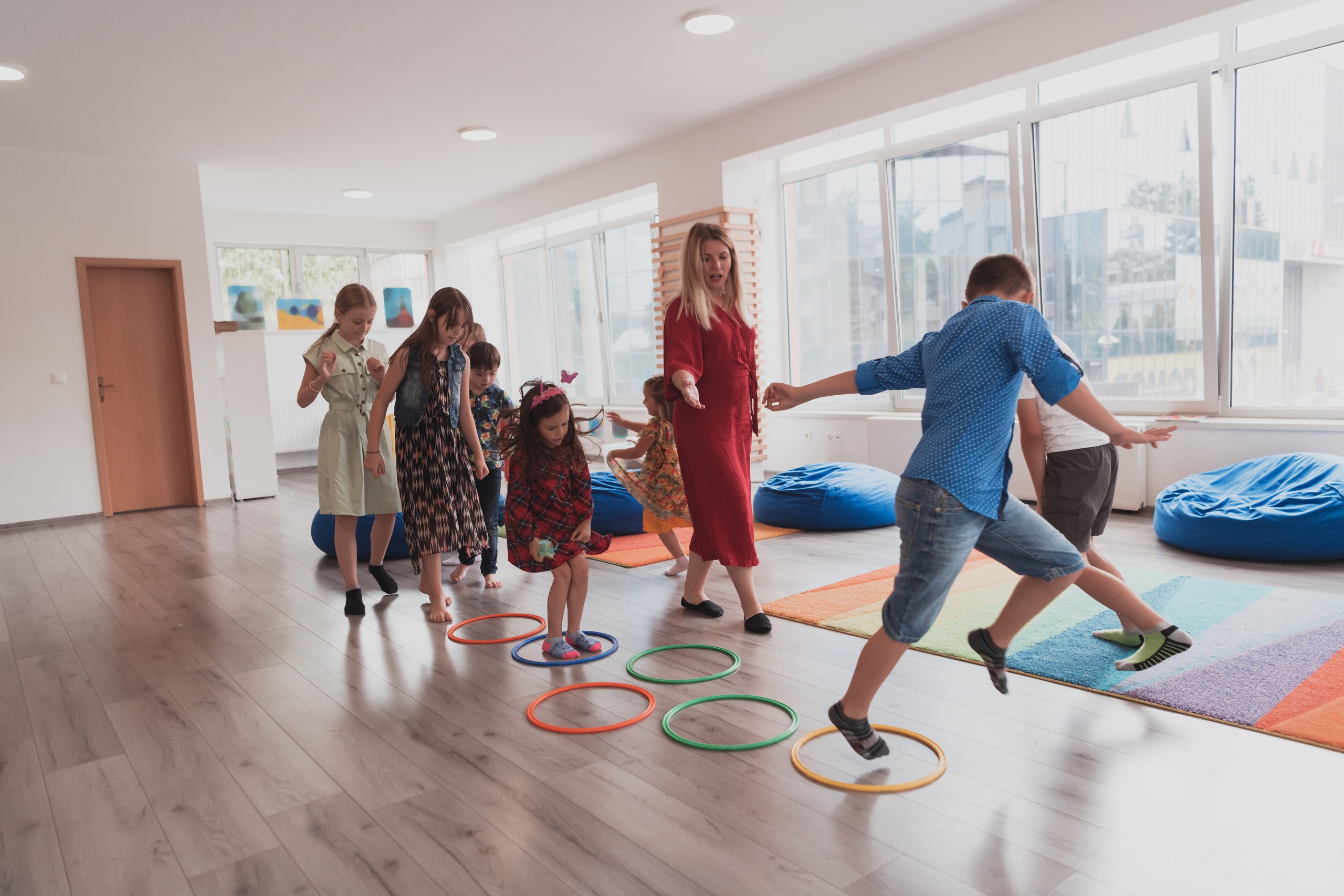 Small nursery school children with female teacher on floor indoors in classroom, doing exercise. Jumping over hula hoop circles track on the floor.
