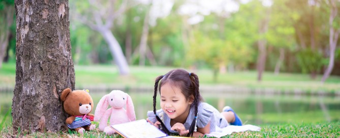 Photo of a girl reading a book outdoors laying on the grass.